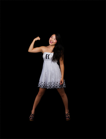 Attractive Japanese American Woman Standing In Dress Showing Bicep Muscels In One Arm On Black Background