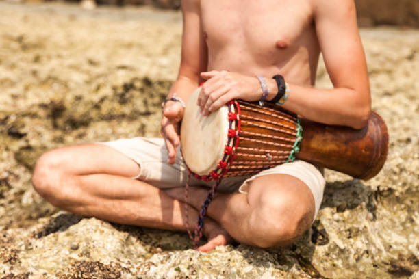 Close up young man drummer, playing rhythm with djembe drum, sitting at tropical coral reef coast background. Travel vacation and music relaxation concept. Selective focus. Copy text space