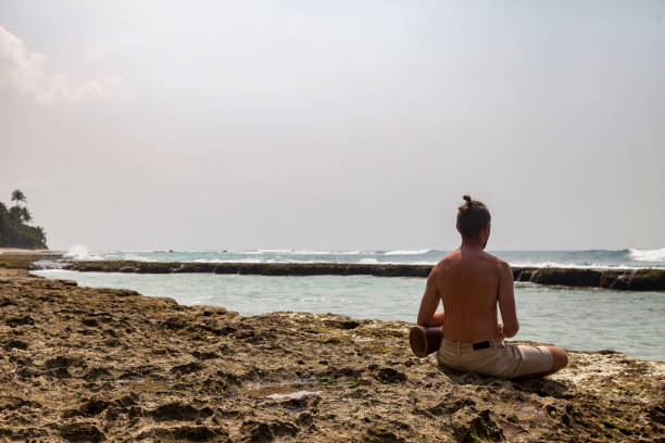Rear view young man drummer, playing rhythm with djembe drum, sitting on tropical coral reef coast at ocean background. Travel vacation and music relaxation concept. Selective focus. Copy text space vista trasera del joven baterista, tocando el ritmo con el tambor djembe, sentado en la costa de arrecifes de coral tropicales en el fondo del océano. concepto de vacaciones de viaje y relajación musical. enfoque selectivo. copiar espacio de texto