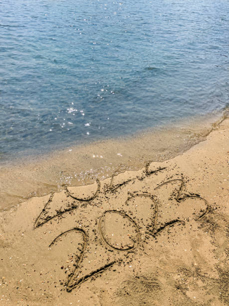 Stock photo showing elevated view of the year dates 2022 and 2023 written in the sand on a sunny beach by the sea's water's edge at low tide with threat of being wiped away. New Year's in southern hemisphere concept. imagen en primer plano de escritura dibujada en una playa soleada con fechas de año 2022 y 2023 escritas en arena con palo, borde del mar durante la marea baja, números escritos a mano en arena dorada suave, vista elevada, concepto de año nuevo en el he