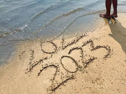 Stock photo showing elevated view of the year dates 2022 and 2023 written in the sand on a sunny beach by the sea's water's edge at low tide with threat of being wiped away. New Year's in southern hemisphere concept.