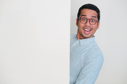 Adult Asian man hiding behind blank white billboard. Peeking out with excited expression