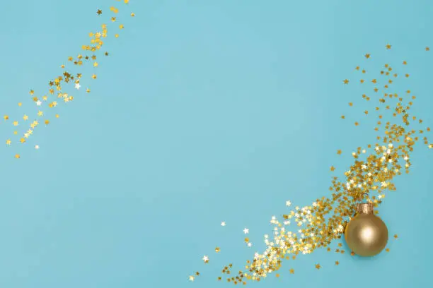 Matte golden Christmas ball and star shaped shining confetti on light blue background. Sparkle sequins. Copy space. Flat lay