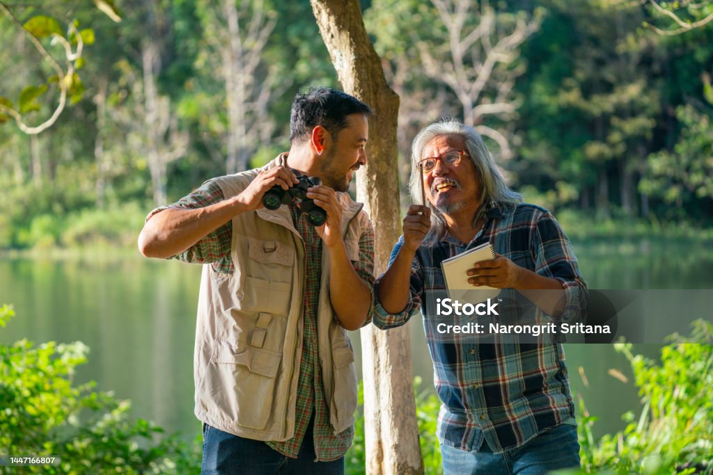 Front view of young Asian man hold binoculars and discuss with senior man who hold book that record some data and they look happy with some new finding. Bird Watching Stock Photo
