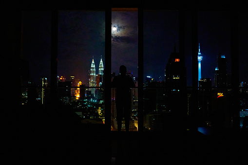 Silhouette of a man on a balcony enjoying the night landscape of downtown Kuala Lumpur. View of the famous petronas oil company towers. Kuala Lumpur, Malaysia - 06.05.2020