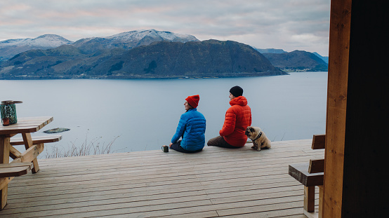 Aerial view of woman and man contemplatoing a view of the scenic fjord during sunset relaxing in the shelter in Norway