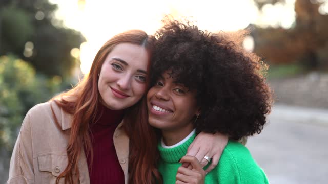 Portrait of a young lesbian couple posing together in front of the camera , Multiracial homosexual women look at each other with a smile,