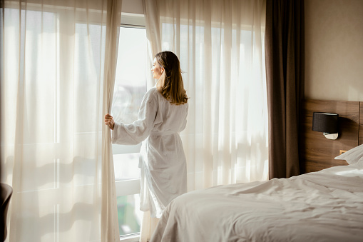 Woman enjoys magnificent view from window in modern apartment or hotel. Young female wearing white bathrobe opening curtains in the morning