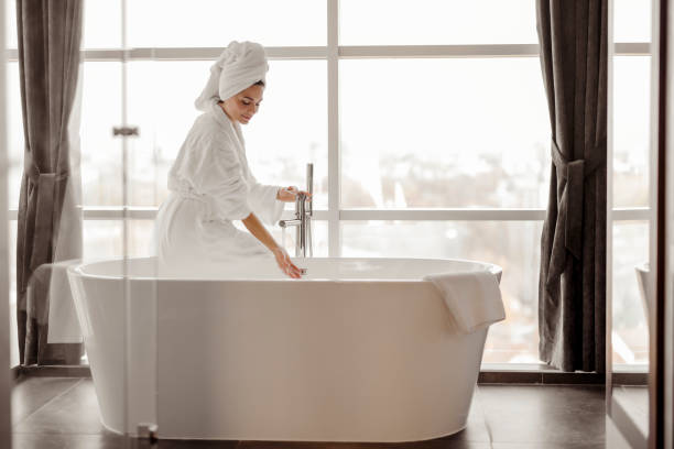 Happiness is a long relaxing bath Woman in luxurious hotel bathroom letting water in the bathtub. Woman wearing white robe playing with water flowing from tap guest bathroom stock pictures, royalty-free photos & images