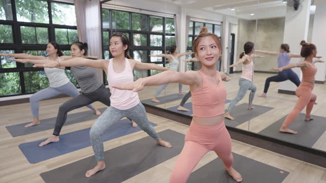 Group of female yoga students doing Warrior I and Warrior II  continuously in front of big mirror in an exercise room of a fitness center with smiley faces.