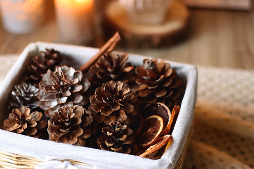 Basket with pine cones and seasonal spices on the table. Lit candles in the background. Hygge at home. Selective focus.