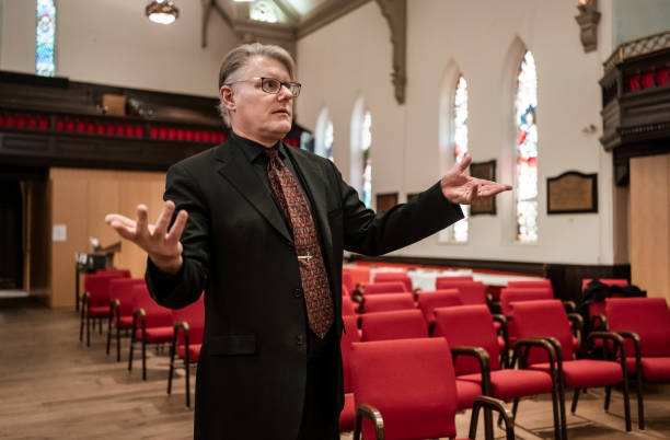 A mature black man wearing church choir robes, standing in front of a red curtain with a serious expression.
