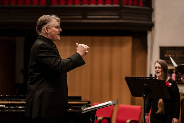 Mature man Church Choir Conductor during Concert performance. He is dressed in all black suit and tie, wearing eyeglasses. Interior of Anglican Church at night.