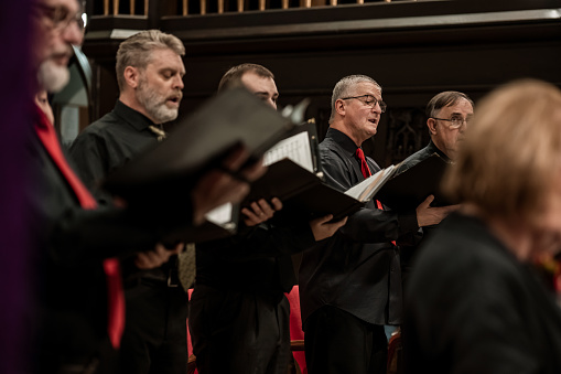 Close ups of men singers in Church Choir during performance at Concert during Christmas Holiday season. Mixed age group of people dressed in all black attire. Interior of Anglican church at night.