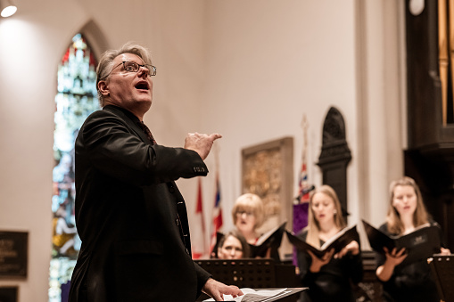 Mature man Church Choir Conductor during Concert performance. Choir in the background. He is dressed in all black suit and tie, wearing eyeglasses. Interior of Anglican Church at night.