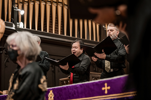 Close ups of men singers in Church Choir during performance at Concert during Christmas Holiday season. Mixed age group of people dressed in all black attire. Interior of Anglican church at night.