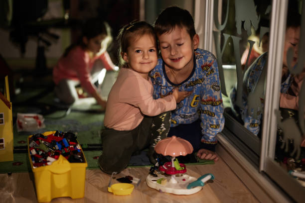 Two Asian children enjoy a sunny day outdoors, sitting in lush grass. The boy smiles at the camera while the girl plays with small objects, sharing a playful family moment in nature.