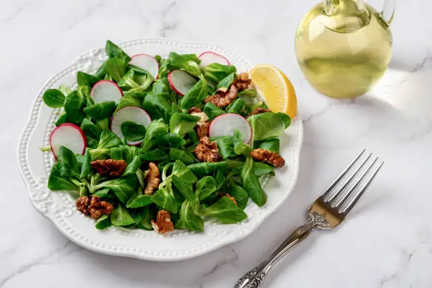 Lambs lettuce salad with radish and walnuts. Cornsalad leaves, nut kernels and sliced radish on a white plate against marble surface. Low calories slimming diet with vitamin vegetable salad. Front view.