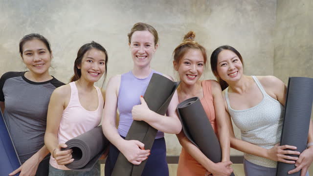 Young women standing against grey wall, holding yoga mats and walking toward camera while talking to each other enjoyably. All of them are looking at camera with big smile.
