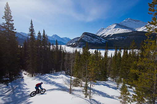 A man goes for a winter fat bike ride in the Rocky Mountains of Canada. Fat bikes are mountain bikes with oversized wheels and tires for riding on the snow. He wears a ski helmet and goggles, and warm winter clothing.