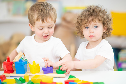 Two young children sit at a desk as they play with colorful blocks together.  They are both dressed casually in white t-shirts as they build and use their imaginations.