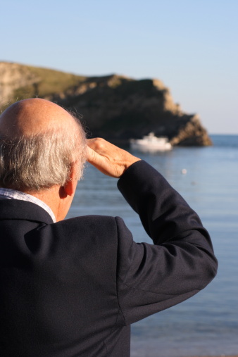 Senior retired man watching a small boat coming into harbour whilst covering his eyes or maybe a salute.
