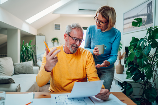 Senior married couple is actively discussing repairs in a new apartment or house while measuring the dimensions in the blueprints and sitting at the table and making notes. Investing into new property