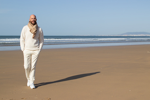 Handsome bald man in scarf strolling on beach. Confident fashionable middle aged bearded man in sweater holding hands in pockets while walking alone. Vacation concept
