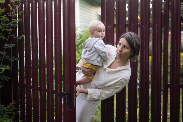 neugierige frau mit einem baby späht das tor des zauns auf - fence child neighbor peeking stock-fotos und bilder