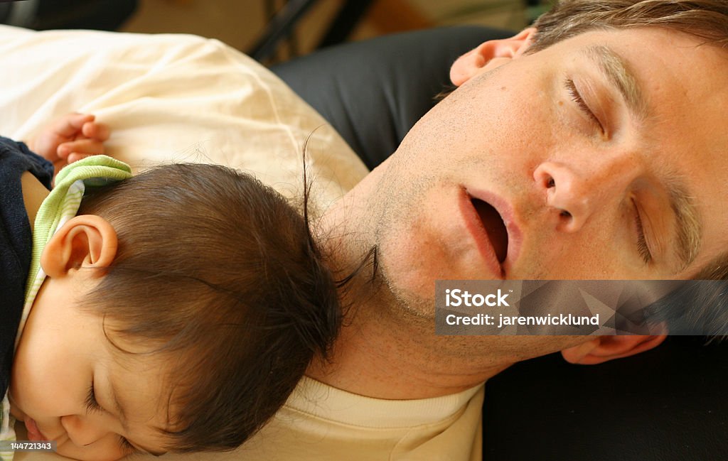 Baby boy asleep on chest of sleeping father Baby boy asleep on chest of sleeping father, while reclining in living room. Adult Stock Photo
