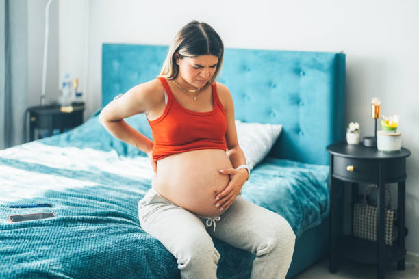 Pregnant young woman sitting on the bed experiencing pain in the lower back Pregnant woman feeling pain while sitting on bed First Symptoms of Pregnancy stock pictures, royalty-free photos & images