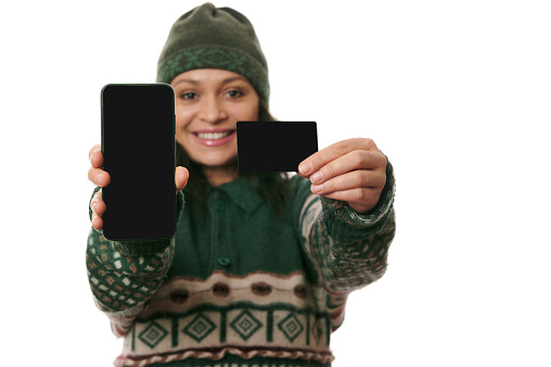 Selective focus on black blank credit card and smartphone with copy ad space in the hands of pretty woman in warm winter clothes, smiling a toothy smile looking at camera, isolated on white background
