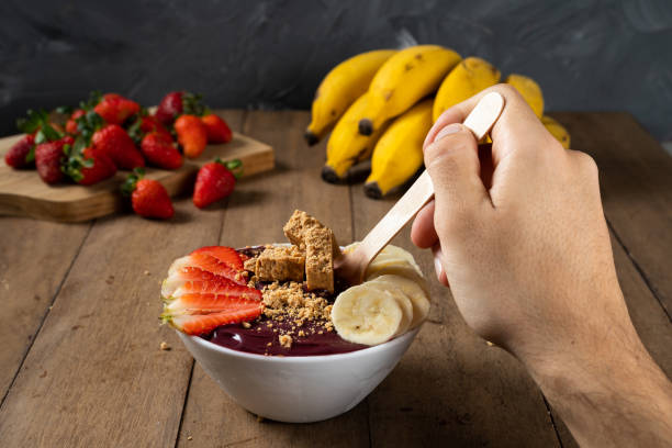 cucchiaio e una ciotola bianca di bacche di açai congelate brasiliane con paçoca, fragola e banana. su una scrivania di legno - men banana holding cut out foto e immagini stock