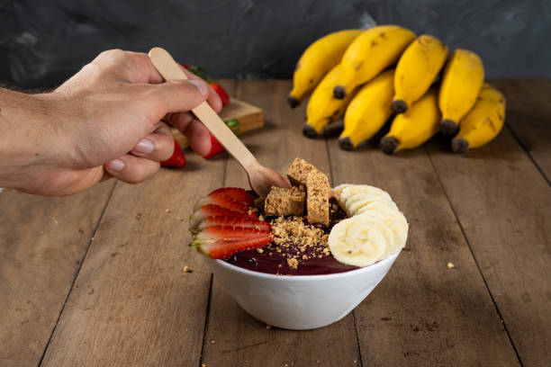 cucchiaio e una ciotola bianca di bacche di açai congelate brasiliane con paçoca, fragola e banana. su una scrivania di legno - men banana holding cut out foto e immagini stock