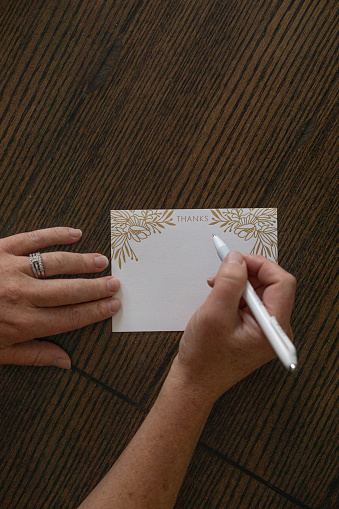 Hands in frame writing a thank you note with gold accent detail on a dark wooden desk