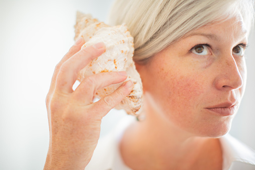 Woman against a light airy background listening into a conch shell against her ear