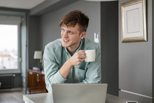 Portrait of young man in shirt leaning on the table while drinking a coffee and using laptop. Home working. Freelance working. E-learning.