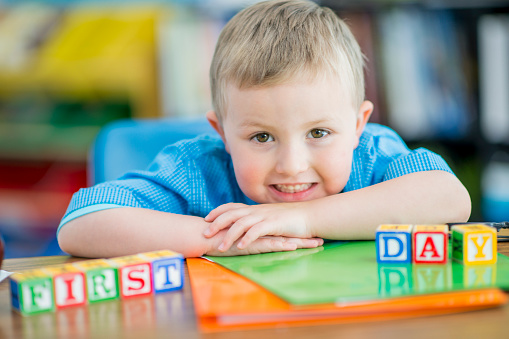 A sweet little blond haired boy sits at his desk with notebooks and colorful letter blocks spread out in front of him.  The blocks spell out 