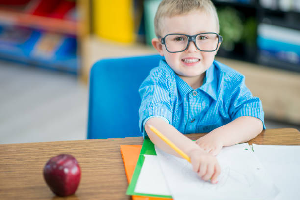 A sweet little blond haired boy with glasses sits at his desk on the first day of school as he works away.  He is dressed casually and has notebooks spread out on the table in front of him.