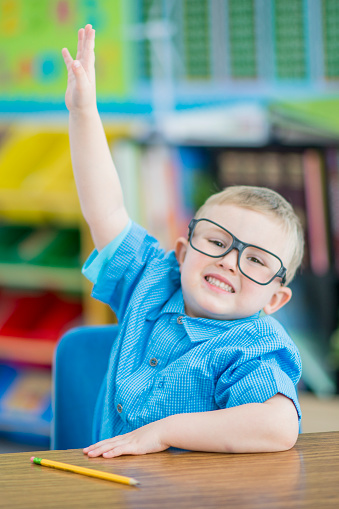 A sweet little blond haired boy sits at a desk on the first day of school as he raises his hand to ask a question.  He is dressed casually and has glasses on as he smiles with enthusiasm.