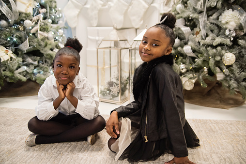 Cute little girls portrait in Christmas setting. They are friends from black ethnicity with pretty black and white party dresses and are looking at the camera with a soft smile. Horizontal full length indoors shot with copy space. This was taken in Montreal, Quebec, Canada.