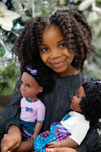 Cute little girl playing with doll that has the same skin colour in Christmas setting. She is black with a pretty black dress and is sitting on the floor in front of the Christmas tree. Vertical waist up indoors shot with copy space. This was taken in Montreal, Quebec, Canada.u