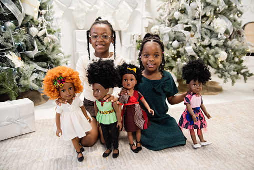 Black little girls playing with same skin colour dolls in Christmas setting. They are dressed with pretty party dresses and are sitting in front of Christmas tree. Horizontal full length indoors shot with copy space. This was taken in Montreal, Quebec, Canada.