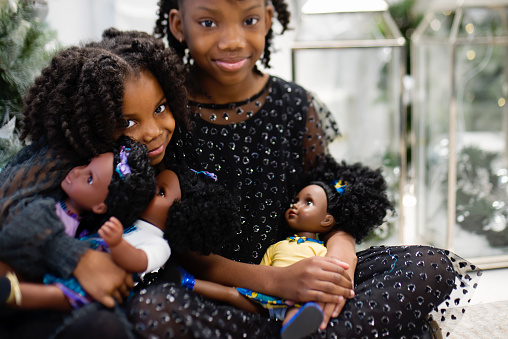 Black little girls playing with same skin colour dolls in Christmas setting. They are sisters with pretty black party dresses and are sitting in front of Christmas tree. Horizontal waist up indoors shot with copy space. This was taken in Montreal, Quebec, Canada.