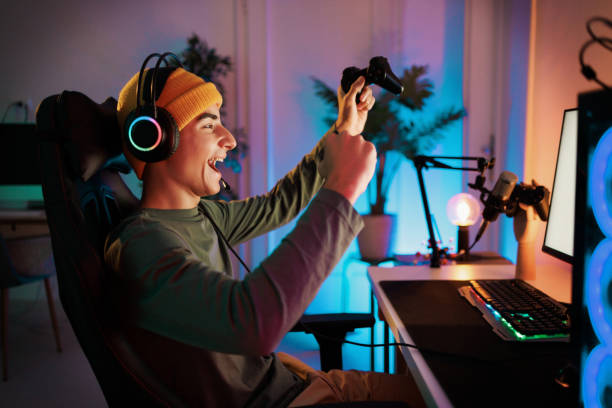 подросток играет за компьютером и празднует победу в видеоигре со сжатым кулаком и улыбкой - gamer стоковые фото и изображения