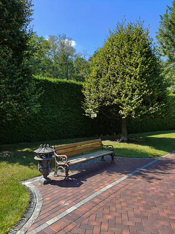 A decorative bench on a tiled sidewalk in a public park against a background of trees and a hedge