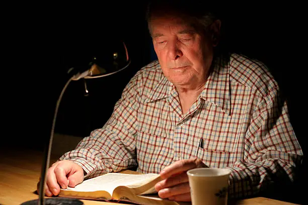 Elderly man reading his KJV Bible by lamplight, while drinking his tea.
