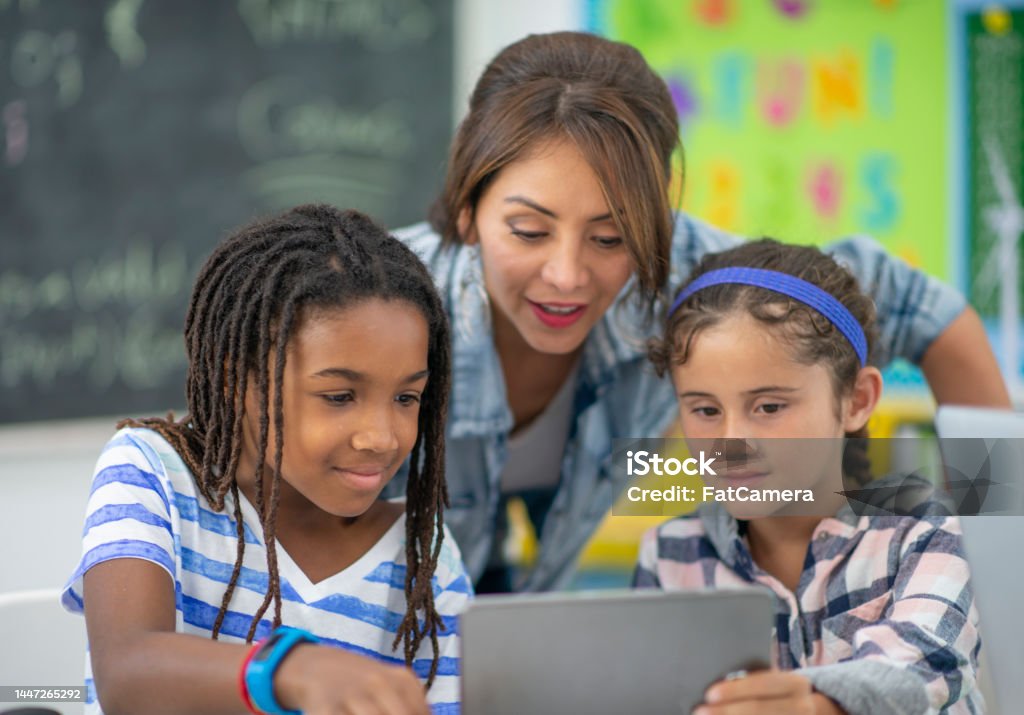 Teacher Assisting Students A female teacher of Hispanic decent, leans in to help two students who are working on a Coding project on a laptop.  The students are dressed casually and are focused on the assignment as they ask the teacher questions. Teacher Stock Photo
