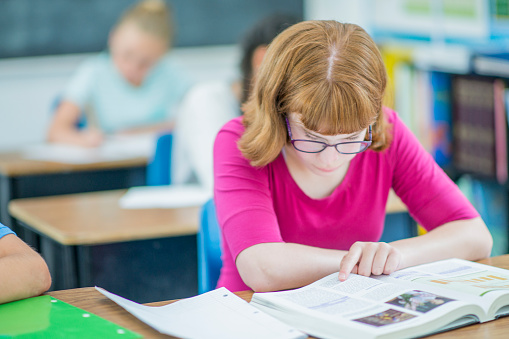 A female elementary student sits at her desk as she works away diligently.  She is dressed casually and has a textbook open in front of her as she focuses on reading the chapter independently.