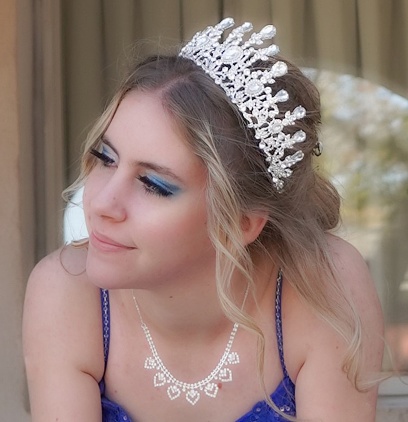 A teen girl, wearing a diamond tiara and diamond heart necklace and royal blue ball gown, looks dreamily off to the side. Close-up of head and shoulders.